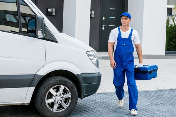 A Locksmith In Blue Overalls Is Carrying A Tool Box And Walking Towards A White Van.